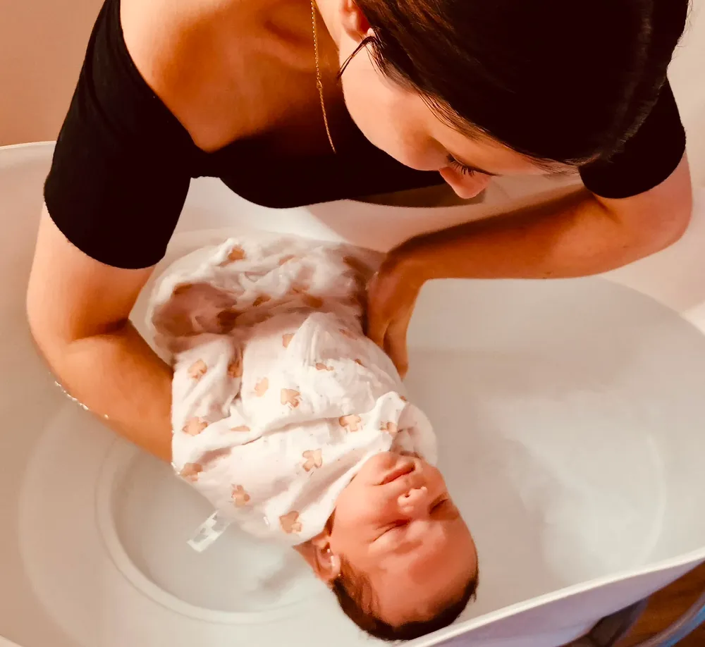 Praticienne accompagnant un bébé emmailloté dans un lange blanc au-dessus du bain
